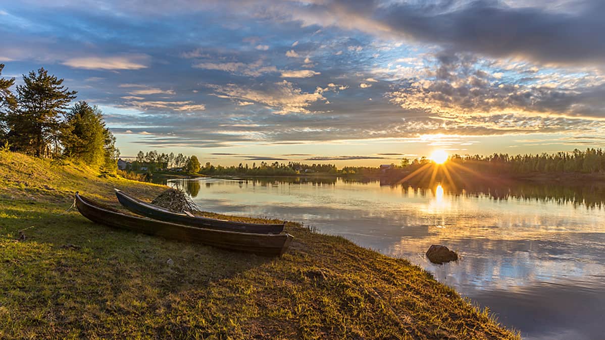 Kuvassa on joen rannalla kaksi venettä ja aurinko paistaa.